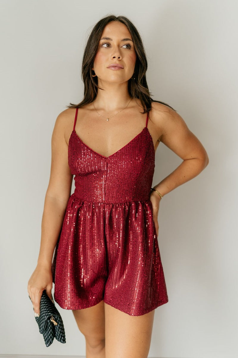 Front view of model wearing the Sasha Burgundy Sequin Strappy Romper that has dark red knit fabric with monochromatic sequins, a v-neckline, adjustable straps, an open back, and a back zipper.