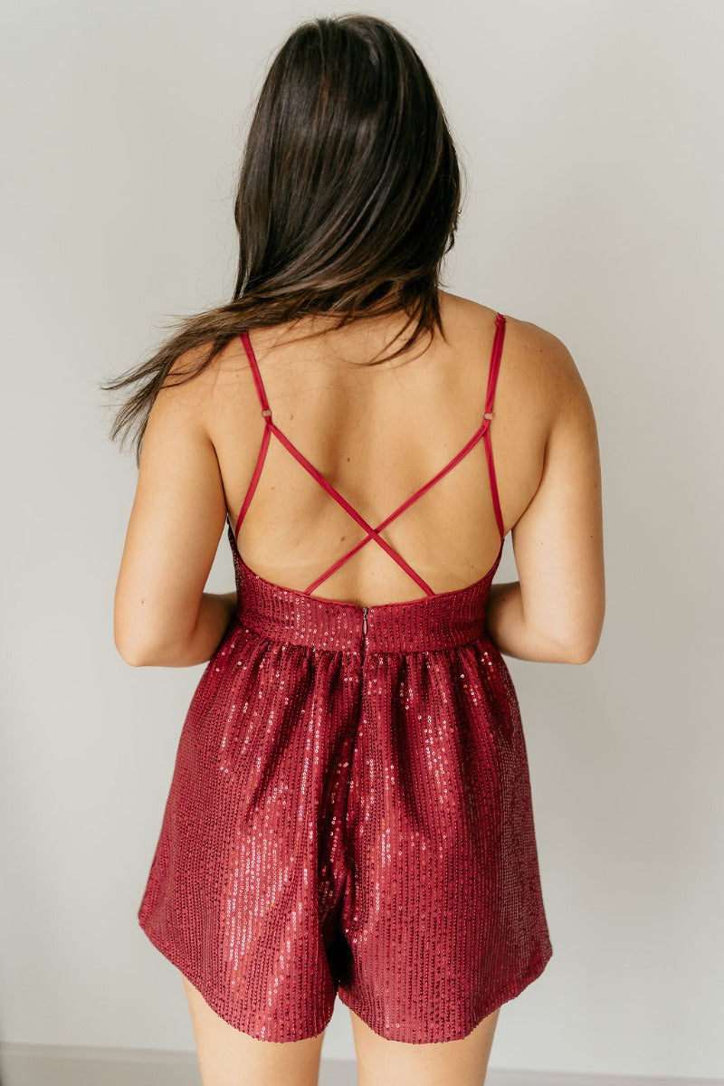 Back view of model wearing the Sasha Burgundy Sequin Strappy Romper that has dark red knit fabric with monochromatic sequins, a v-neckline, adjustable straps, an open back, and a back zipper.