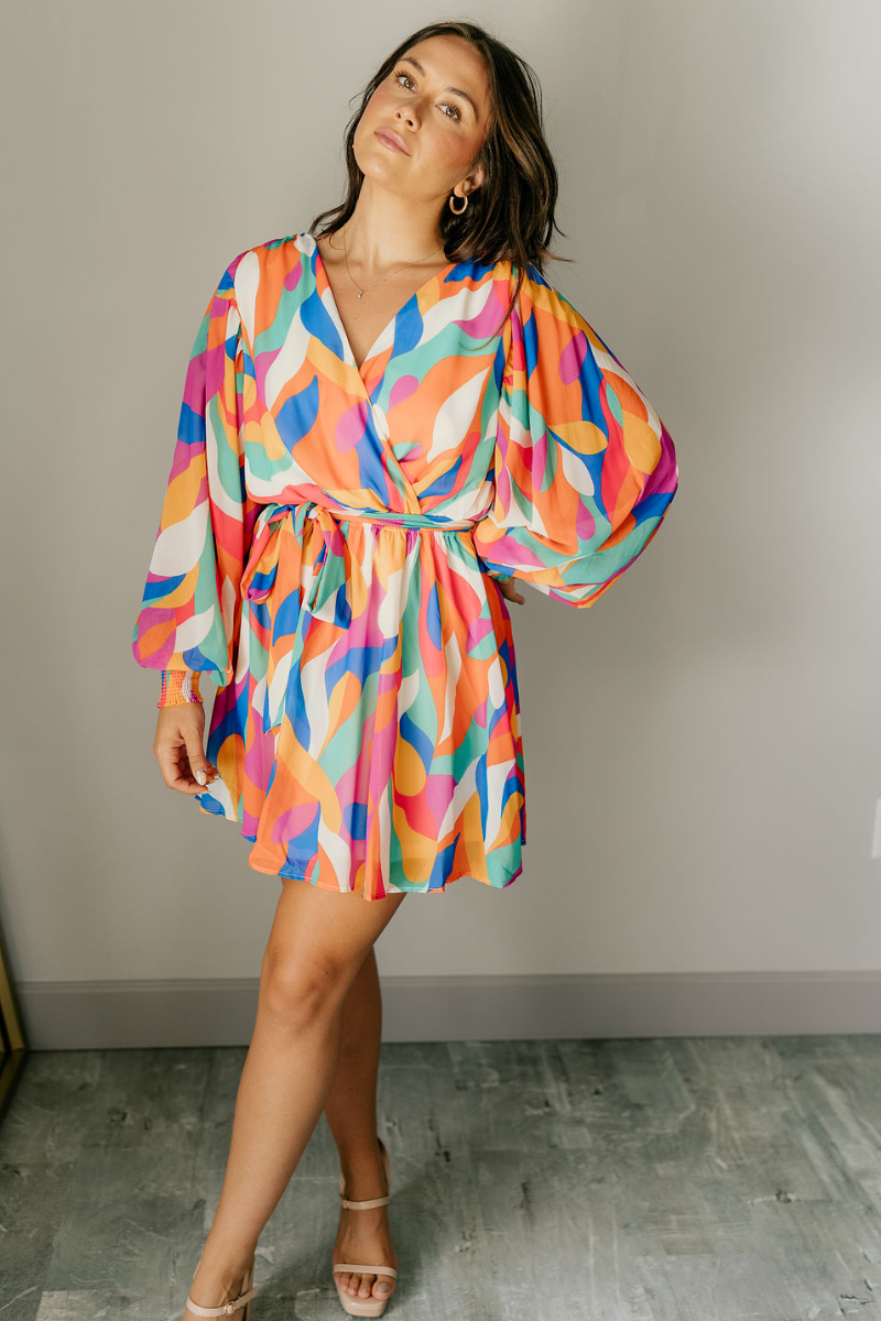 Full body front view of model wearing the Amaya Printed Long Sleeve Mini Dress that has sheer fabric with a colorful swirl pattern, mini length, an elastic waist, a tie belt, a surplice neckline, and long balloon sleeves.