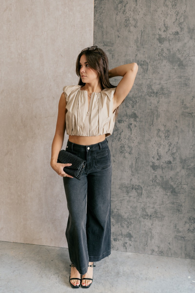 Full body front view of model wearing the Casey Khaki Zip-Front Cropped Tank Top that has khaki textured fabric, a cropped elastic waistband, a monochrome front zipper, a round neckline and a sleeveless design.