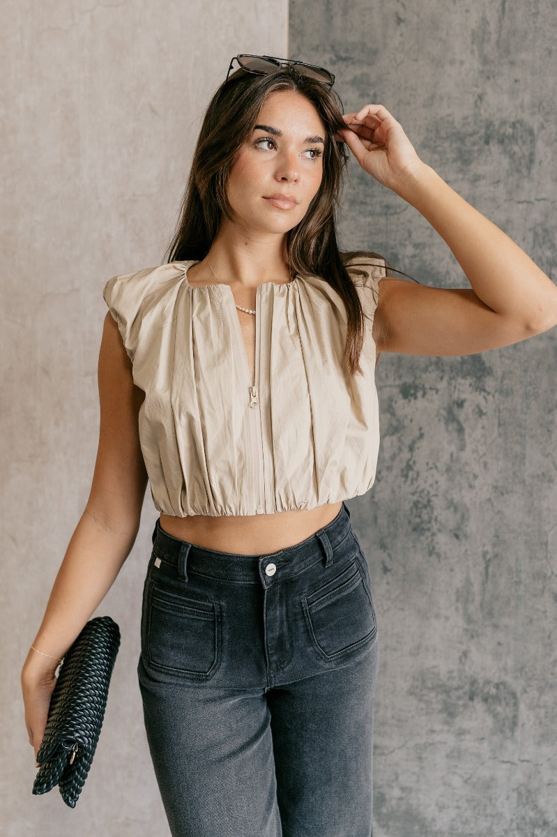 front view of model wearing the Casey Khaki Zip-Front Cropped Tank Top that has khaki textured fabric, a cropped elastic waistband, a monochrome front zipper, a round neckline and a sleeveless design.