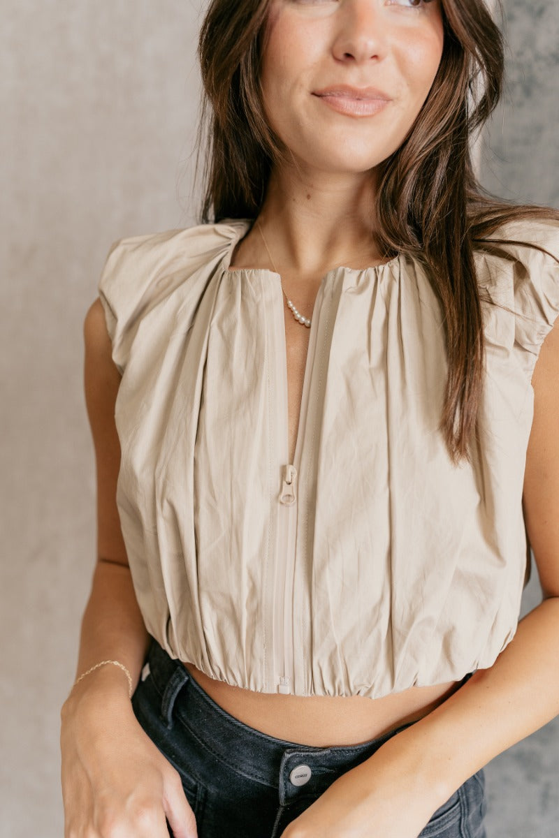Close front view of model wearing the Casey Khaki Zip-Front Cropped Tank Top that has khaki textured fabric, a cropped elastic waistband, a monochrome front zipper, a round neckline and a sleeveless design.