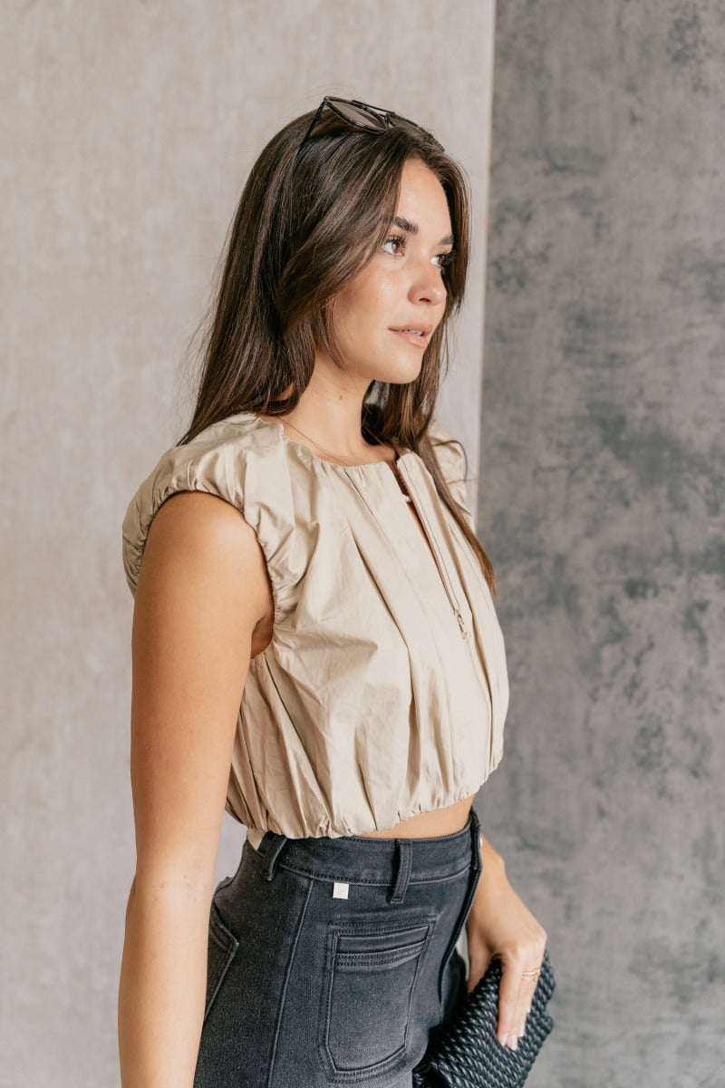 Side view of model wearing the Casey Khaki Zip-Front Cropped Tank Top that has khaki textured fabric, a cropped elastic waistband, a monochrome front zipper, a round neckline and a sleeveless design.