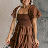 Frontal side view of model wearing the Willow Brown Puff Sleeve Mini Dress which features brown fabric, mini length, two tiered baby doll style, brown lining, a smocked chest, a square neckline and short puff sleeves.