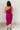 Full body back view of model wearing the Tiffany Fuchsia Ruched One-Shoulder Midi Dress that has fuchsia stretchy semi-satin fabric, ruched details, a one-shoulder neckline, and a side zipper.