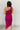 Full body back view of model wearing the Tiffany Fuchsia Ruched One-Shoulder Midi Dress that has fuchsia stretchy semi-satin fabric, ruched details, a one-shoulder neckline, and a side zipper.