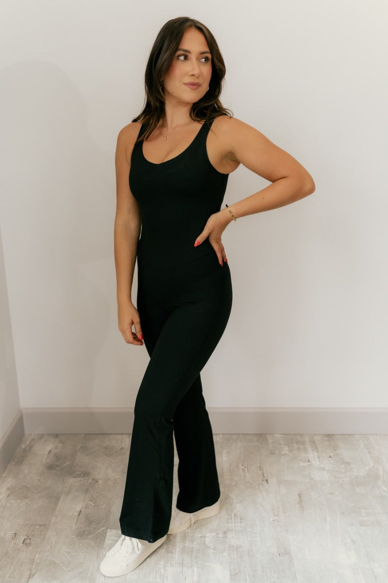Full body front view of model wearing the Yasmin Black Athletic Onesie Jumpsuit that has black athleisure fabric, monochrome stitched details, a scooped neckline, thick straps, and flared pant legs.