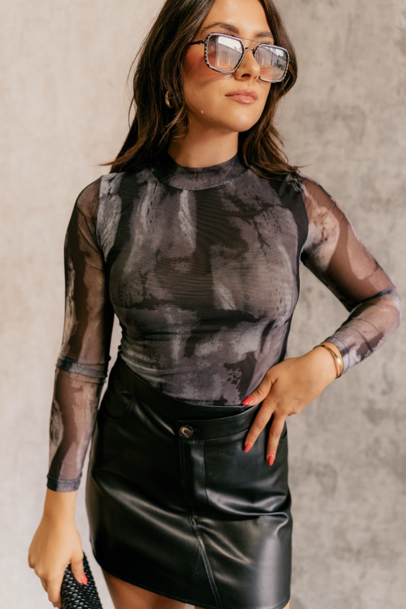 front view of model wearing the Leanna Black Marbled Mesh Long Sleeve Bodysuit that has black, grey and white mesh fabric with a watercolor design, a high neck, long sleeves, and a thong bottom.