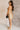 Full body side view of model wearing the Amber Gold Cowl Neckline Sleeveless Mini Dress which features gold shimmer fabric, mini length, gold lining, a cowl neckline, adjustable straps, and a monochromatic back zipper with a hook closure.
