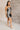 Full body side view of model wearing the Amber Gunmetal Cowl Neckline Sleeveless Mini Dress which features gunmetal grey shimmer fabric, mini length, black lining, a cowl neckline, adjustable straps, and a monochromatic back zipper with a hook closure.
