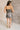 Full body back view of model wearing the Amber Gunmetal Cowl Neckline Sleeveless Mini Dress which features gunmetal grey shimmer fabric, mini length, black lining, a cowl neckline, adjustable straps, and a monochromatic back zipper with a hook closure.