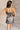 Back view of model wearing the Amber Gunmetal Cowl Neckline Sleeveless Mini Dress which features gunmetal grey shimmer fabric, mini length, black lining, a cowl neckline, adjustable straps, and a monochromatic back zipper with a hook closure.
