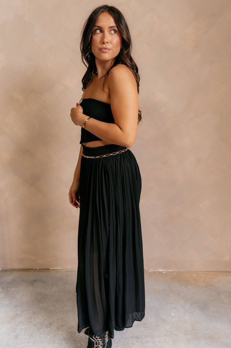 Full body side view of model wearing the Mila Black Pleated Midi Skirt which features black pleated sheer fabric, black lining, midi length, and an elastic waistband with ruffle details.