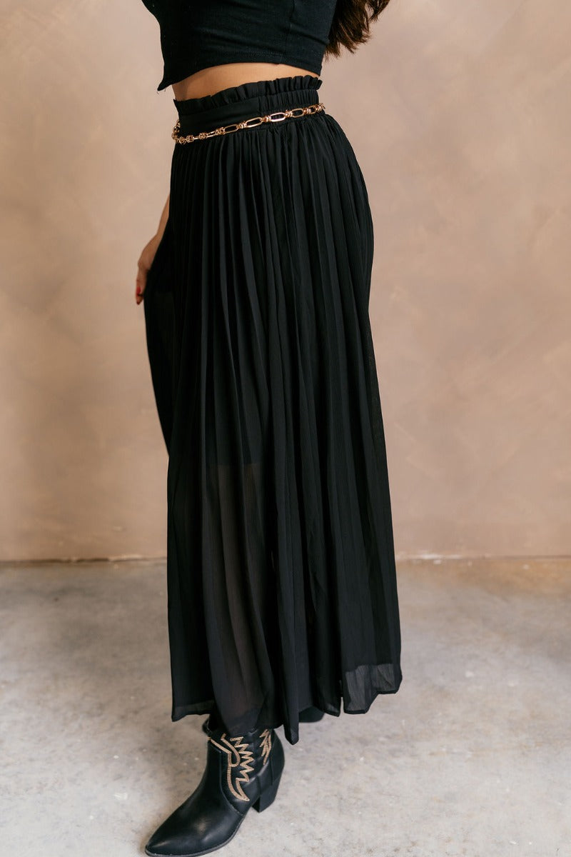 Side view of model wearing the Mila Black Pleated Midi Skirt which features black pleated sheer fabric, black lining, midi length, and an elastic waistband with ruffle details.