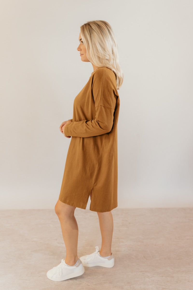 Full body side view of model wearing the Sara Mustard Long Sleeve T-Shirt Dress that has light brown cotton fabric, mini length, slits on each side, a round neckline, dropped shoulders, and long sleeves.