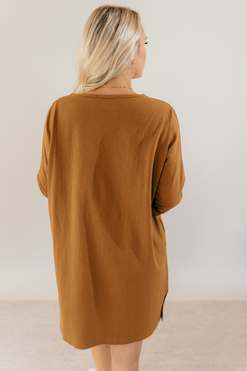 back view of model wearing the Sara Mustard Long Sleeve T-Shirt Dress that has light brown cotton fabric, mini length, slits on each side, a round neckline, dropped shoulders, and long sleeves.