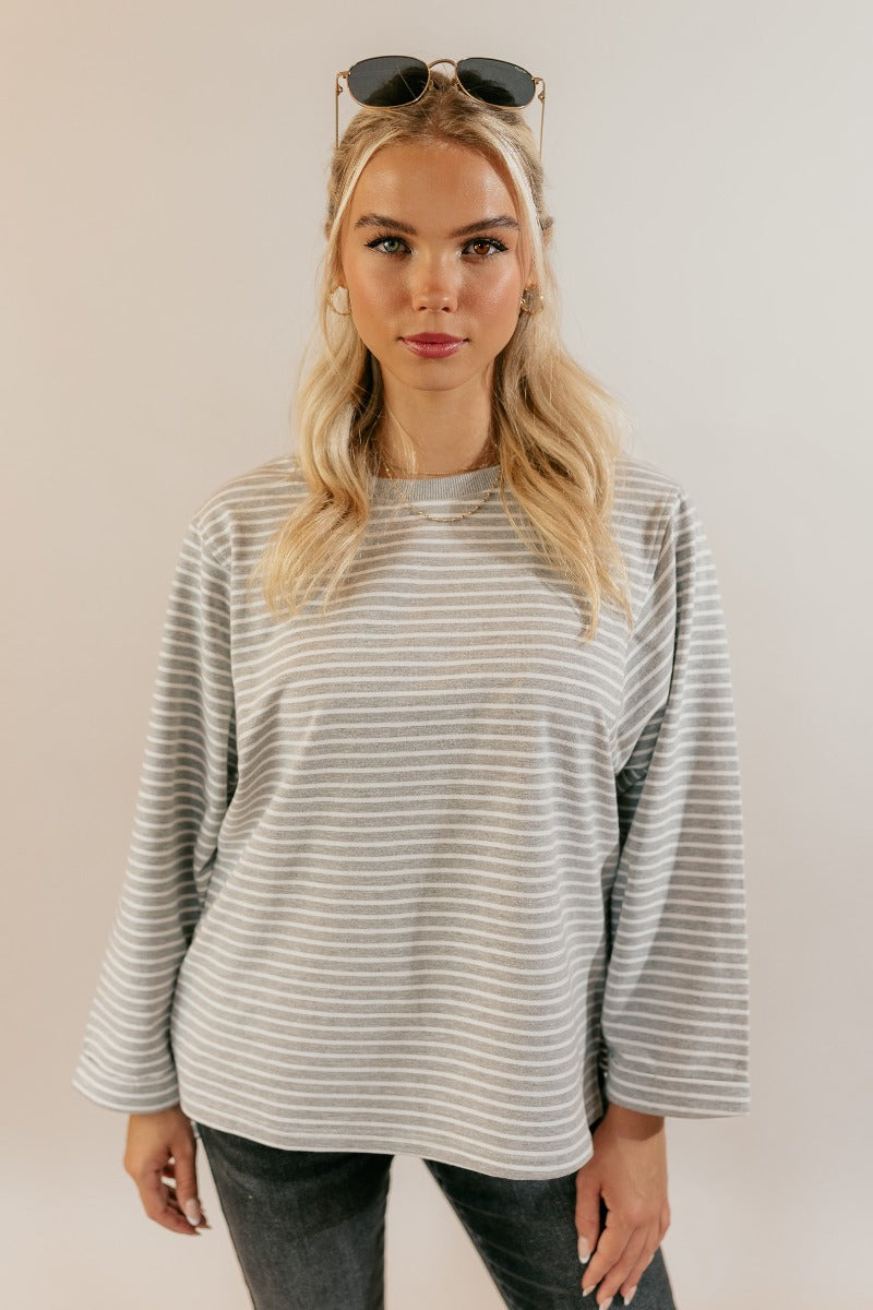 Front untucked view of model wearing the Samantha Grey & White Striped Long Sleeve Top that has grey and white knit fabric, stripe pattern, slits, a round neck, and long flare sleeves.