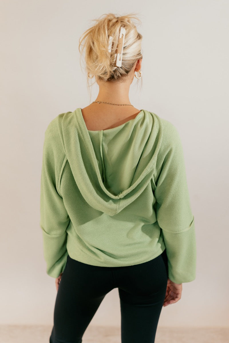 Back view of model wearing the Leila Sage Green Hoodie Sweater that has sage textured fabric, a thick hem, a v-neckline with a hood and drawstring ties, and long puff sleeves with cuffs.
