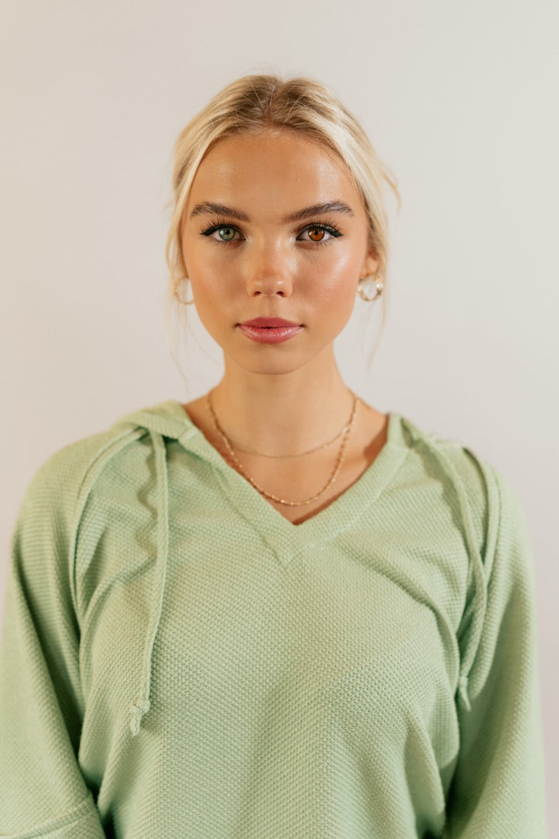 Upper front view of model wearing the Leila Sage Green Hoodie Sweater that has sage textured fabric, a thick hem, a v-neckline with a hood and drawstring ties, and long puff sleeves with cuffs.