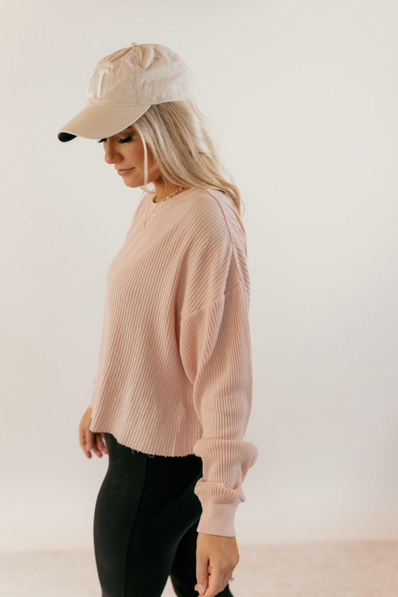 Side view of model wearing the Ada Blush Waffle-Knit Long Sleeve Top that has light pink textured fabric, a raw hem, a round neckline, dropped shoulders, and long sleeves with cuffs.