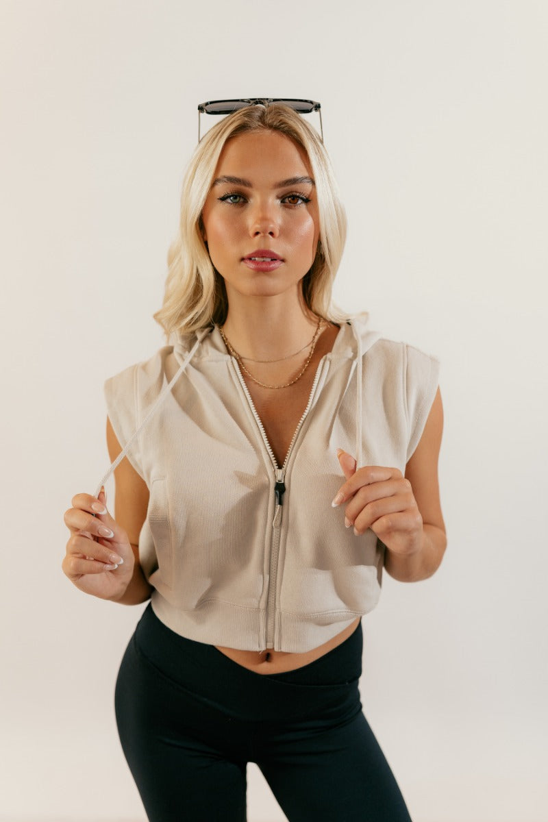 front view of model wearing the Melanie Taupe Sleeveless Cropped Hoodie that has taupe knit fabric, a cropped waist, pockets, a front zipper a hood, and a sleeveless design.