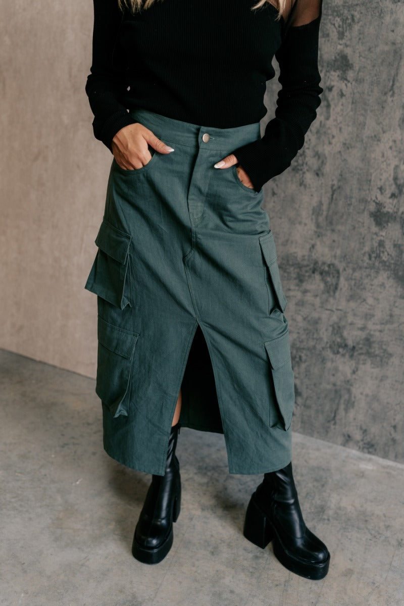 front view of model wearing the Ember Green Front Slit Cargo Midi Skirt that has green fabric, a front slit, pockets, a front zipper with a button closure, and a back elastic waistband.