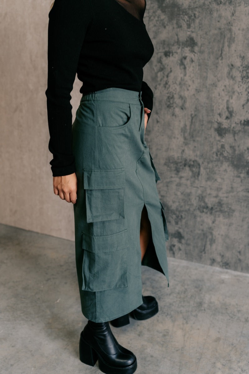 Side view of model wearing the Ember Green Front Slit Cargo Midi Skirt that has green fabric, a front slit, pockets, a front zipper with a button closure, and a back elastic waistband.