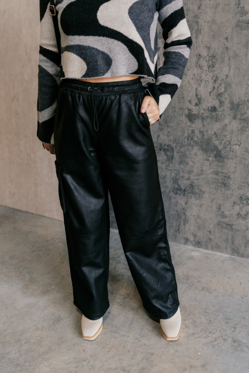 front view of model wearing the Zuri Black Faux-Leather Pants that have black soft faux leather fabric, two front pockets, an elastic waistband with a drawstring tie, and wide legs.
