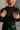 Close front view of model wearing the Anastasia Black Embellished Mesh Top that has black mesh fabric, silver embellishments, exposed seam details, a high neckline, and long sleeves.