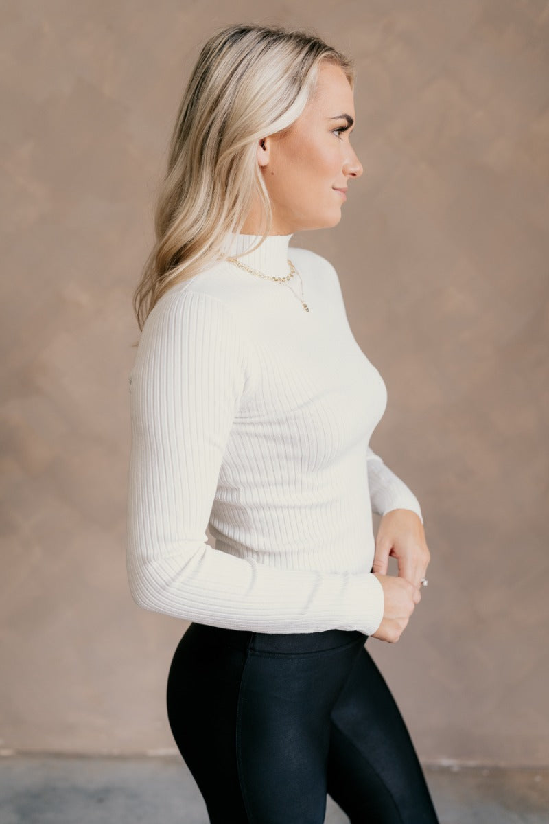 side view of model wearing the Norah Off White Mock Neck Top that has off white ribbed fabric, a high neckline, and long sleeves.