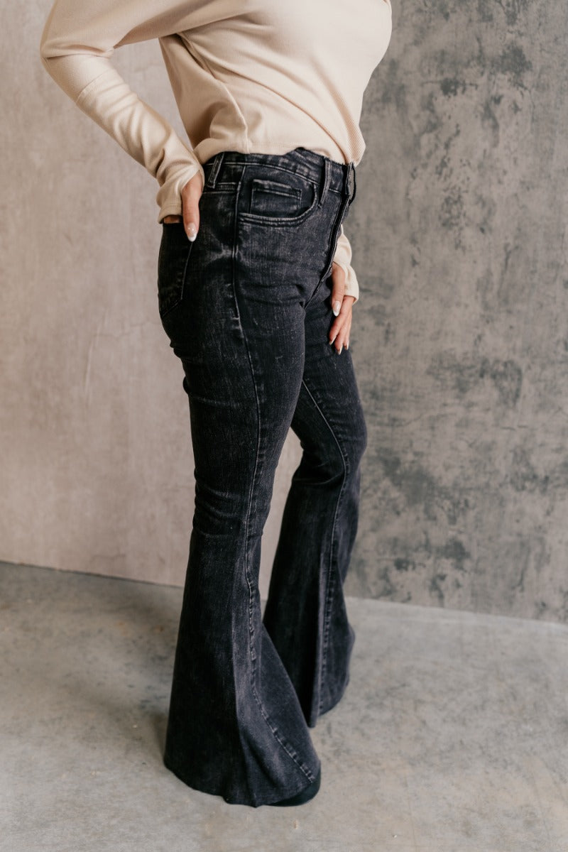 Side view of model wearing the Rooted Denim: Kamila Black Flare Jeans that have washed black denim fabric, a button-up fly, belt loops, two front pockets, two back pockets and flared legs.