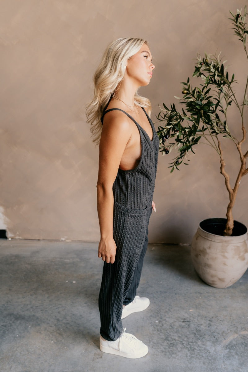 Full body side view of model wearing the Presley Charcoal Knit Sleeveless Jumpsuit that has charcoal grey textured knit fabric, two side pockets, a v-neckline, adjustable straps, and jogger pant legs.