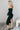 Full body side view of model wearing the Hayley Black Ribbed Off-The-Shoulder Long Sleeve Midi Dress which features black knit fabric, midi length, a slit in the back, a square neckline, and long sleeves.