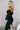 Side view of model wearing the Hayley Black Ribbed Off-The-Shoulder Long Sleeve Midi Dress which features black knit fabric, midi length, a slit in the back, a square neckline, and long sleeves.