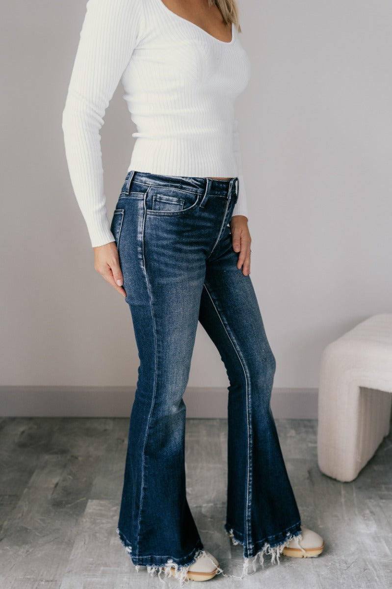 Full body side view of model wearing the Rooted Denim: Leah Dark Wash Flare Leg Jeans which features dark wash denim fabric, a front zipper with a button closure, two front pockets, two back pockets, belt loops, and flared legs with fray hem.