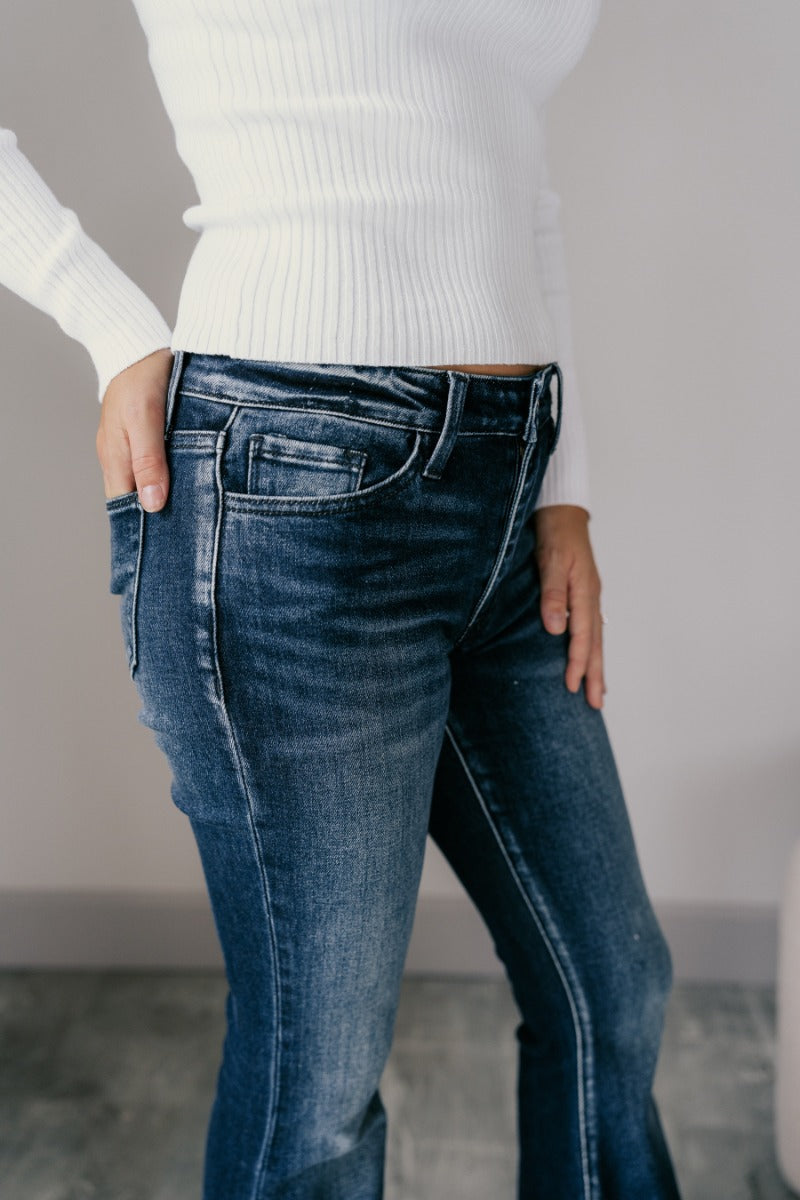 Close up side view of model wearing the Rooted Denim: Leah Dark Wash Flare Leg Jeans which features dark wash denim fabric, a front zipper with a button closure, two front pockets, two back pockets, belt loops, and flared legs with fray hem.