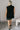 Full body back view of model wearing the Wren Black Sleeveless Turtleneck Sweater which features black knit fabric, a high-low hem, slits on each side, a turtleneck neckline, and a sleeveless design.