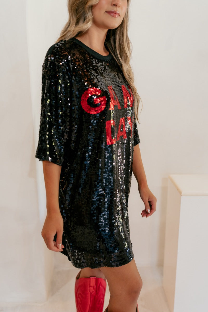 Side view of model wearing the Game Day Red & Black Sequin Dress that has black sequins, red sequins that say "Game Day", and black knit back, an oversized fit, short sleeves, and a round neck.