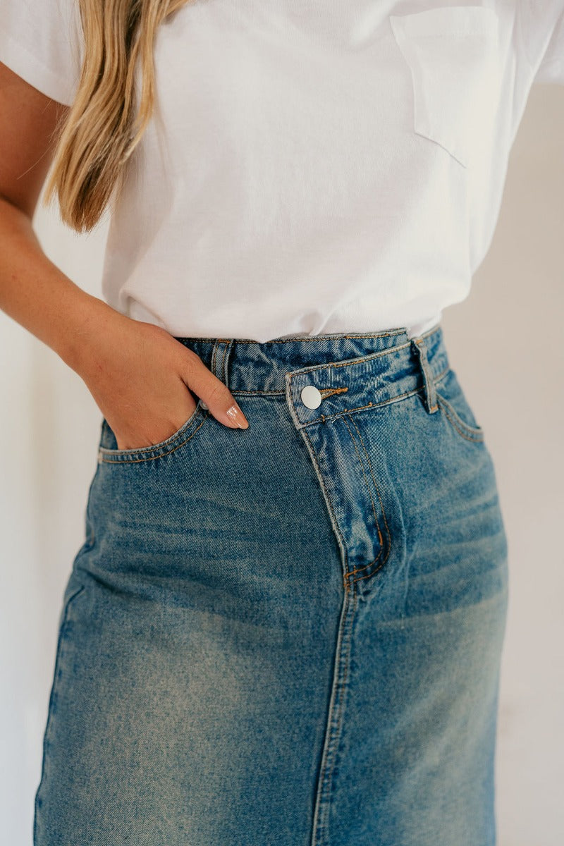 Close up view of model wearing the Skye Blue Denim Slit Midi Skirt which features washed blue denim fabric, a front slit, a front slanted zipper with a button closure, belt loops, front pockets, back pockets, and maxi length.