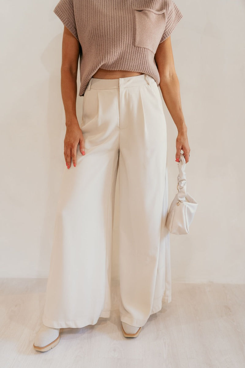 front view of model wearing the Scarlett Beige Wide Leg Pants that have light beige lightweight fabric, two front pockets, a front zipper with a hook closure, belt loops, and wide legs.