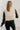 Back view of model wearing the Tahoe Cream Faux Leather Puffer Cropped Vest which features cream faux leather puffer fabric, a cropped waistline with a drawstring, a front zip-up closure, two front pockets, and a high neckline.