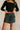 Front view of model wearing the Piana Washed Black Denim Mini Skirt which features washed black denim fabric, mini length, an overlap design with frayed hem, two front snap closures, one front pocket with a snap cloure and an adjustable strap, and a monoc