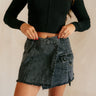 Front view of model wearing the Piana Washed Black Denim Mini Skirt which features washed black denim fabric, mini length, an overlap design with frayed hem, two front snap closures, one front pocket with a snap cloure and an adjustable strap, and a monoc