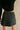 Back view of model wearing the Piana Washed Black Denim Mini Skirt which features washed black denim fabric, mini length, an overlap design with frayed hem, two front snap closures, one front pocket with a snap cloure and an adjustable strap, and a monoch