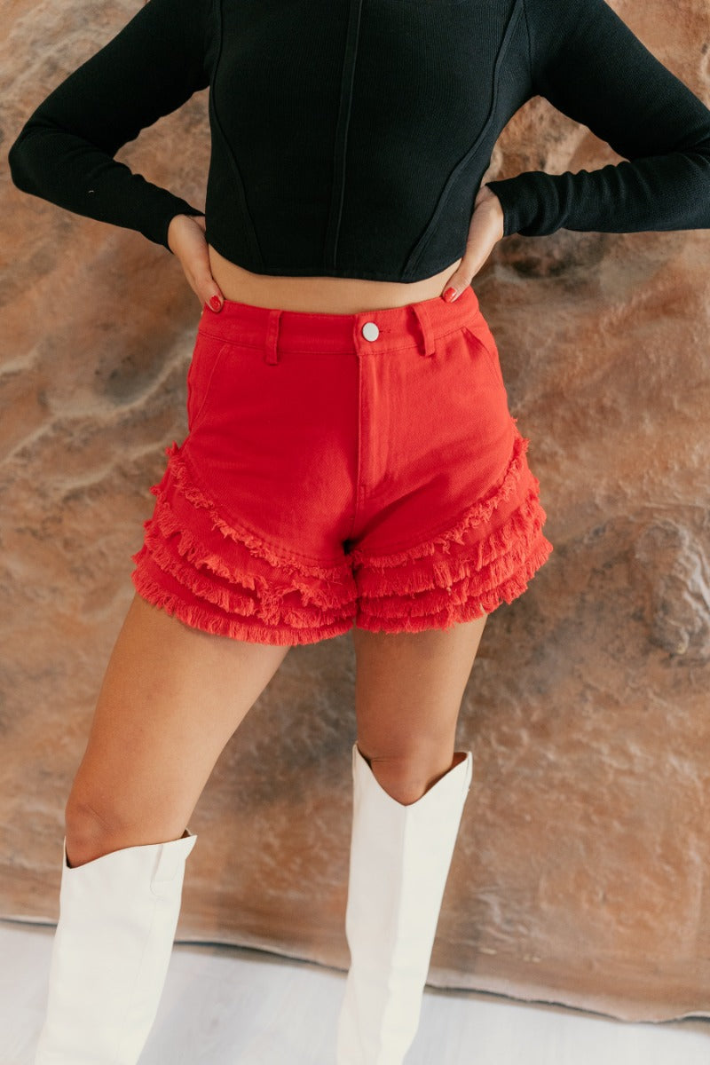 front view of model wearing the Skylar Red Tiered Raw-Edge Denim Shorts that have washed red denim fabric, distressing, pockets, a front zipper with a button, belt loops, and a frayed tiered hem.