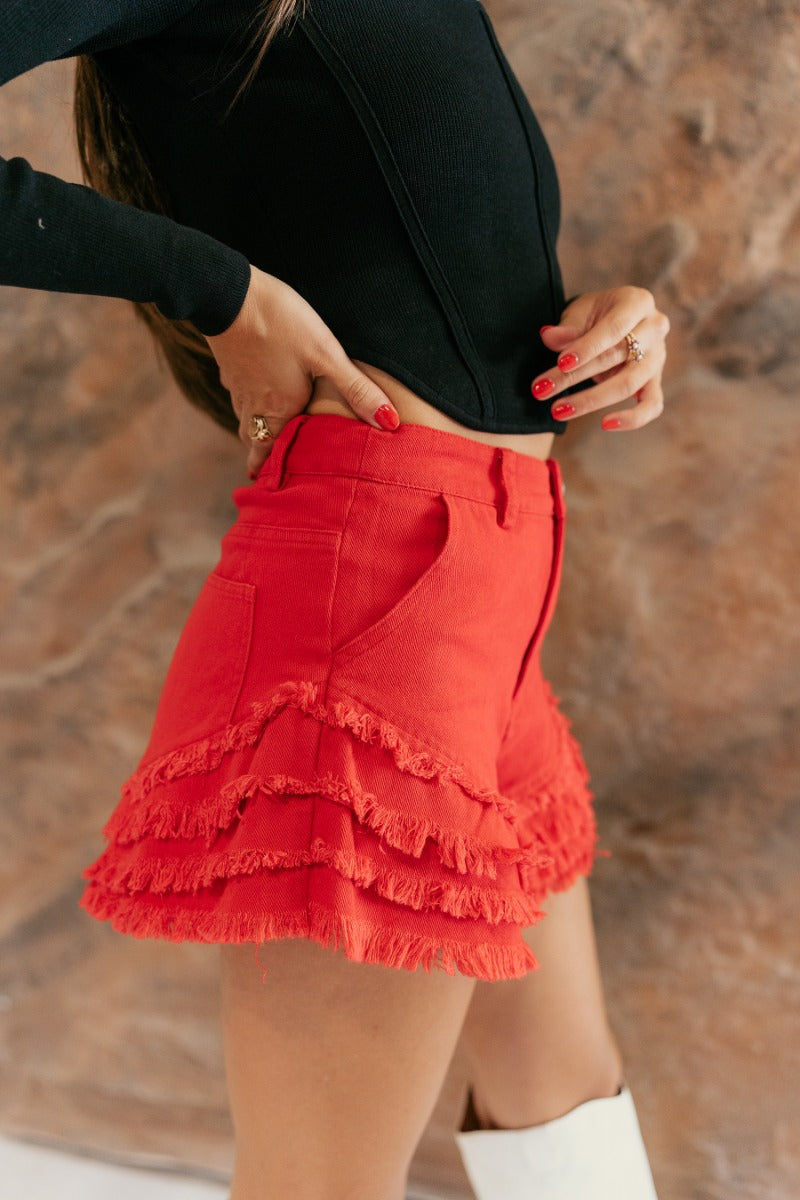 Side view of model wearing the Skylar Red Tiered Raw-Edge Denim Shorts that have washed red denim fabric, distressing, pockets, a front zipper with a button, belt loops, and a frayed tiered hem.
