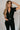 Front view of model wearing the Rielle Black Adjustable Belt Short Sleeve Jumpsuit which features black fabric, two front pockets, a front zipper, an adjustable belt with a silver buckle, a collared neckline, short sleeves, and straight pant legs.