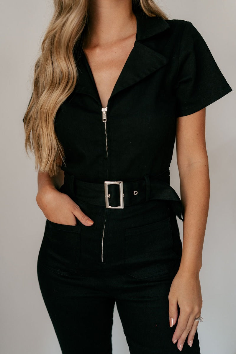 Close up view of model wearing the Rielle Black Adjustable Belt Short Sleeve Jumpsuit which features black fabric, two front pockets, a front zipper, an adjustable belt with a silver buckle, a collared neckline, short sleeves, and straight pant legs.