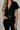 Close up view of model wearing the Rielle Black Adjustable Belt Short Sleeve Jumpsuit which features black fabric, two front pockets, a front zipper, an adjustable belt with a silver buckle, a collared neckline, short sleeves, and straight pant legs.
