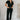 Full body view of model wearing the Rielle Black Adjustable Belt Short Sleeve Jumpsuit which features black fabric, two front pockets, a front zipper, an adjustable belt with a silver buckle, a collared neckline, short sleeves, and straight pant legs.